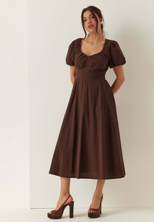 Donna che indossa un vestito midi marrone con maniche a sbuffo e scollatura quadrata, abbinato a sandali marroni con plateau e tacco alto.
