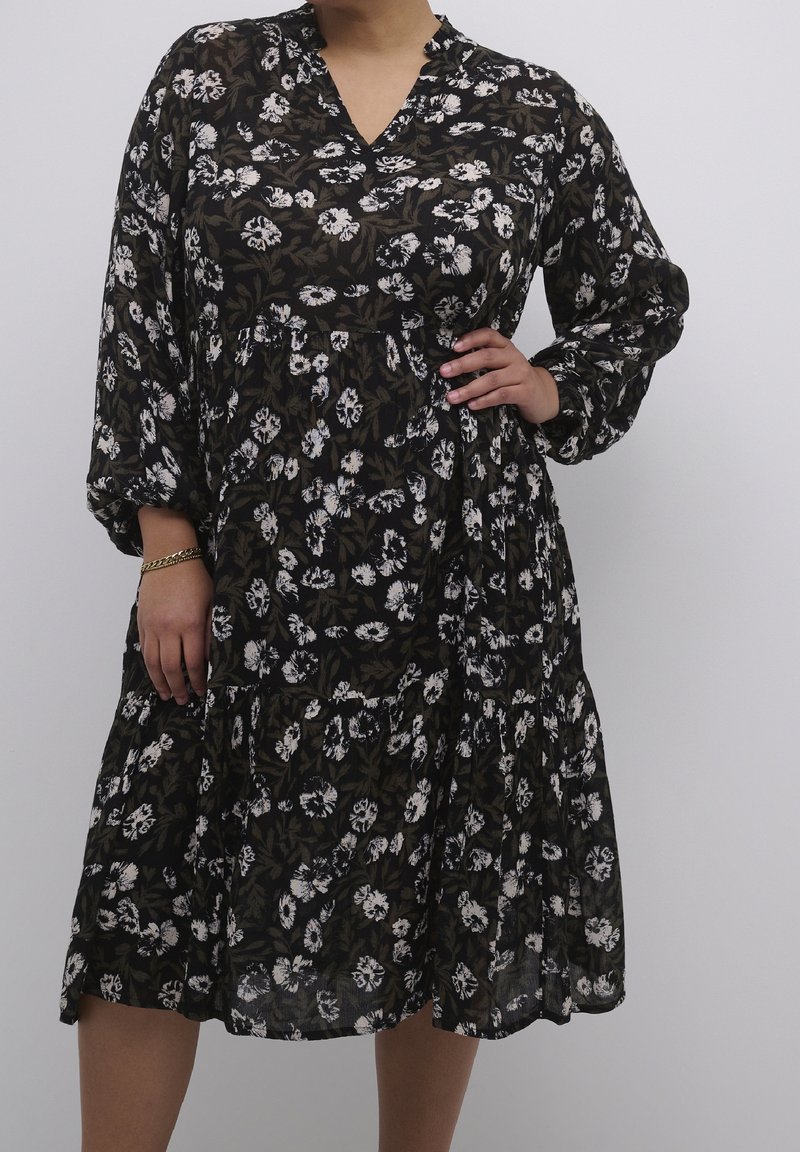 Femme portant une robe longue noire à fleurs avec un décolleté en V, une main sur la hanche et l'autre détendue sur le côté.