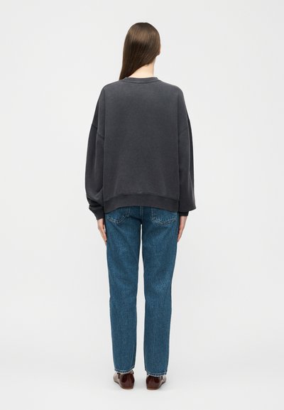 Asmuo ilgais plaukais, dėvintis išblukusį juodą per didelį džemperį, mėlynas tiesias džinsus ir rudus batus, stovintis nugara.