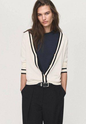 Cremefarbener Cardigan mit schwarzem Besatz, tiefem V-Ausschnitt und Knopfverschluss. Kombiniert mit einem navyfarbenen Rollkragenpullover und hoch taillierten schwarzen Hose.