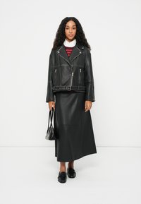 Μαύρη δερμάτινη biker ζακέτα με ζώνη στη μέση, συνδυασμένη με μια μακριά μαύρη φούστα. Το μοντέλο κρατά μια μικρή μαύρη τσάντα. Λευκό γιακά κάτω από ένα κόκκινο ριγέ πουλόβερ.