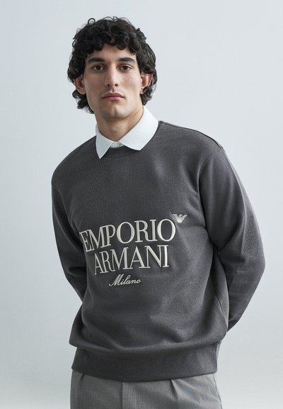 Sweat-shirt gris à texture côtelée, doté d'un grand logo brodé "EMPORIO ARMANI Milano", associé à une chemise blanche à col.