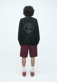 Sudadera negra con un gran logo de árbol gris en la espalda, combinada con pantalones cortos burdeos. Presenta un corte relajado y puños acanalados.