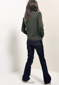 Jeans acampanados de color azul oscuro con costuras blancas contrastantes, combinados con una camiseta de manga larga en verde oliva oscuro, usados con zapatillas blancas.