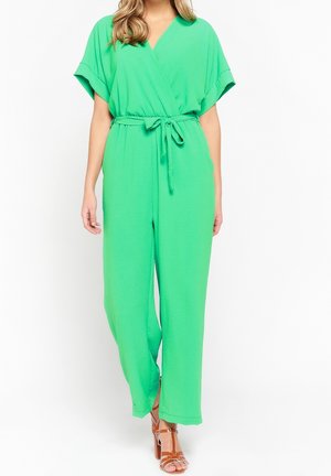 Kobieta ubrana w jasnooliwkowy kombinezon z krótkim rękawem, dekoltem w serek i wiązaniem w talii, w połączeniu z brązowymi sandałami na wysokim obcasie ze paskami.