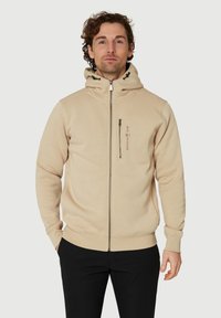 Sudadera con capucha beige, de cuello alto, con cierre frontal y bolsillos laterales. Presenta un logo y cordones de contraste en negro en la capucha.