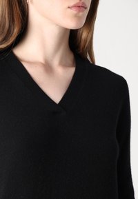 Juodas v-kaklo megztinis, pagamintas iš minkštos medžiagos, su tekstūruotu paviršiumi ir prigludusiu dizainu. Matoma siūlė prie kaklo.