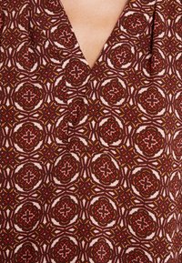 Blusa estampada com um decote em V profundo. O material é um tecido suave com um design geométrico em borgonha, rosa e verde. Detalhes texturizados por toda a peça.