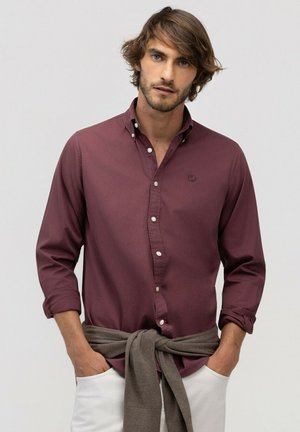 Hombre con barba clara que lleva una camisa burdeos abotonada, pantalones blancos y un suéter gris atado alrededor de la cintura, con las manos en los bolsillos.