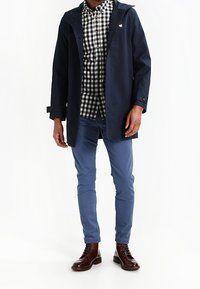 Marinblå vattentät kappa med huva, lagd över en svartvit rutiga skjorta, matchad med ljusblå byxor och bruna kängor.