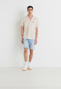 Short-sleeve, cream-colored striped shirt with buttons, paired with light blue denim shorts and white sneakers, standing on a plain background.