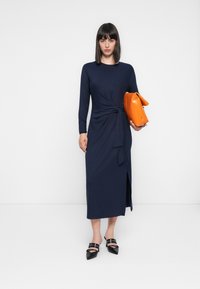 Kvinne med rett, mørkt hår iført en lang marineblå kjole med sideutskjæring, som holder en stor oransje clutch veske og svarte spisse hæler.