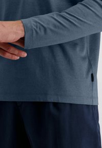 Person, der den Ärmel eines langärmligen blauen Hemdes anpasst, das mit dunkelblauen Hosen getragen wird, und die Stoffstruktur sowie die Nähdetails zeigt.