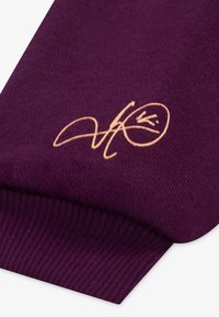 Violetinės megztinio rankovės su raukšlėta tekstūra, turinčios aukso spalvos siuvinėtą logotipą ant audinio.