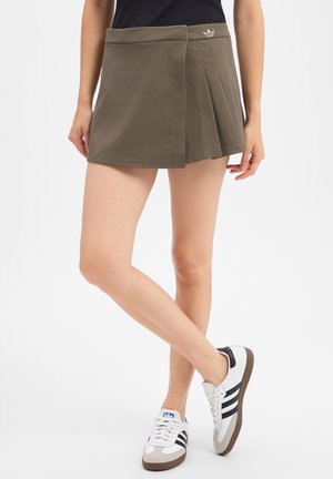 Femme portant une mini-jupe plissée vert olive et des baskets blanches avec des bandes noires, debout sur un fond blanc.