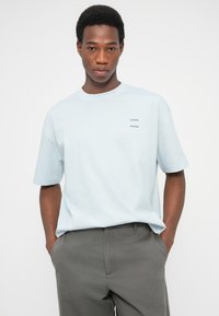 Ανοικτό γαλάζιο βαμβακερό μπλουζάκι με κοντά μανίκια, με δίχρωμο λογότυπο κειμένου στο αριστερό στήθος. Το μοντέλο το συνδύασε με γκρι παντελόνια.