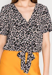 Frau trägt eine schwarze, floral gemusterte Kurzarmbluse mit Knöpfen, die an der Taille gebunden ist, kombiniert mit einem leuchtend gelben Rock.