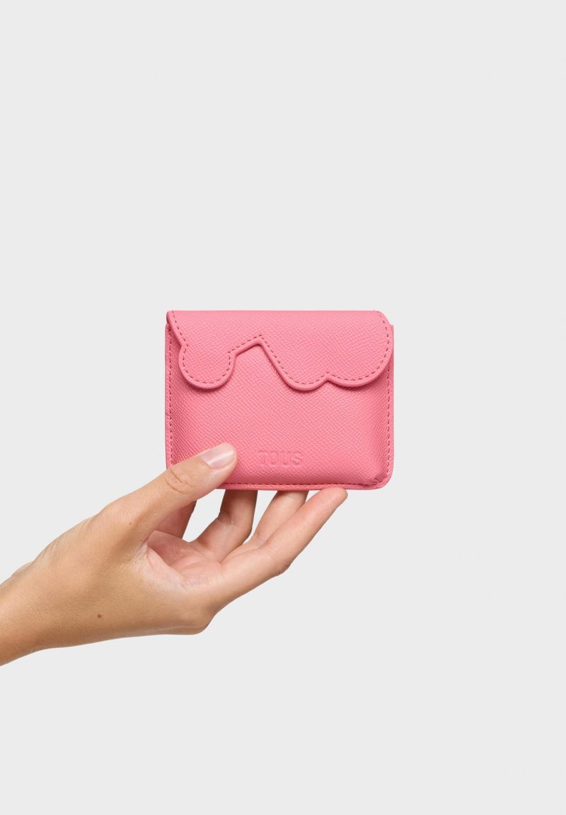 Dłoń trzymająca mały różowy portfel ze skóry z falistą klapką i tłoczonym nazwiskiem marki na gładkim jasnym tle.