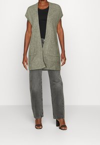 Cardigan verde lavorato a maglia con apertura frontale, maniche a cappuccio e due tasche frontali, abbinato a un top nero e jeans grigi a vita alta.