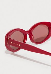 Red sunglasses with octagonal frames, shaded lenses, and a textured red arm. Features gold hardware and engraved logo on the temple.