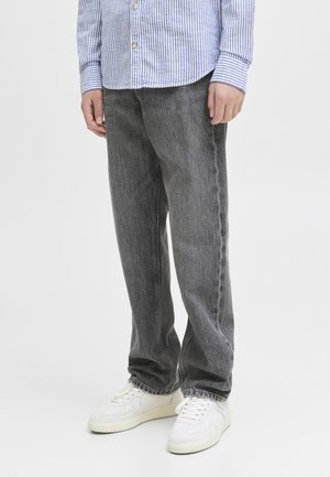 Personne portant un jean gris foncé ample, des baskets blanches et une chemise rayée bleue et blanche, debout devant un fond uni.