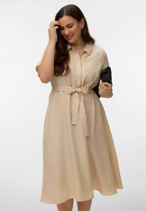 Femme souriante, portant une robe beige ceinturée, tenant une pochette noire sous un bras et touchant ses cheveux de l'autre main.