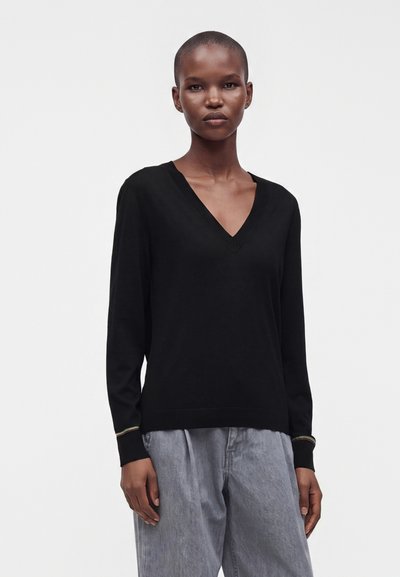 Personne aux cheveux courts portant un haut noir à col en V à manches longues et un pantalon gris ample sur un fond blanc uni.