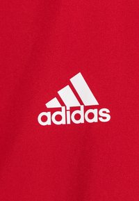 Rode stof met een gladde textuur, met een prominent wit adidas-logo. Het logo bevat drie diagonale strepen en kleine letters.
