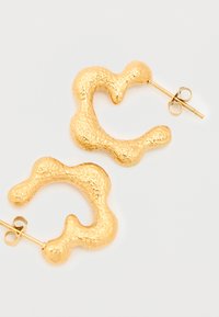 Σκουλαρίκια κρίκοι με κυματιστή υφή σε χρυσό τόνο, με καρφάκι, τοποθετημένα σε απλό ανοιχτό φόντο.