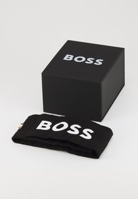Caja negra con logo blanco "BOSS" y accesorio de tela negra doblada que presenta el mismo logo en blanco. Textura suave, diseño minimalista.