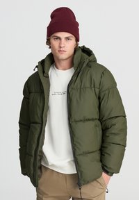 Oliwkowa puchowa kurtka z kapturem, zapinana na zamek błyskawiczny, z elastycznymi mankietami, noszona na jasnym swetrze. Tekstura jest gładka i puszysta.