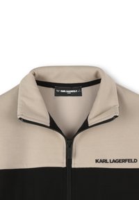 Veste en polaire zippée avec une partie supérieure beige et une partie inférieure noire. Col montant, fermeture éclair visible et étiquette de marque à l'intérieur du col.