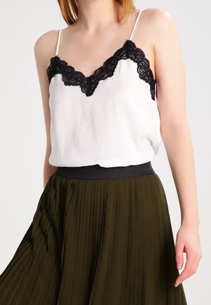 Kvinde iført en hvid undertrøje med sort blonderkant og et højtaljet, plisseret mørkegrønt skørt mod en ensfarvet baggrund.