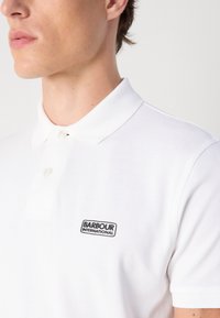 Fehér pólóing, texturált anyagból készült, klasszikus gallérral, két elülső gombbal és fekete Barbour International logóval a mellkason.