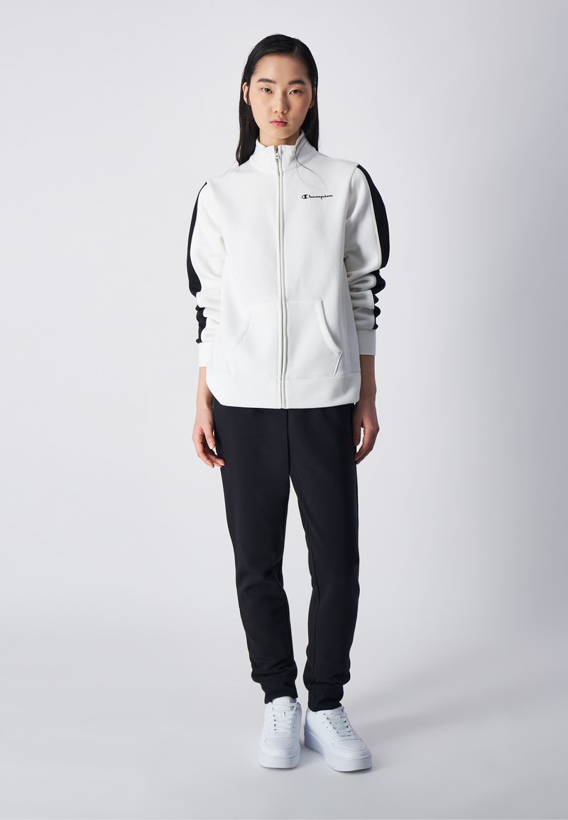 Witte zip-up sweatshirt met zwarte accenten op de mouwen, gecombineerd met zwarte jogger pants en witte sneakers. Kenmerkend logo op de borst.