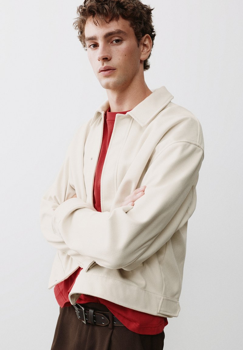 Jeune homme aux cheveux bruns bouclés portant une veste beige par-dessus une chemise rouge et un pantalon foncé, debout les bras croisés devant un fond uni.