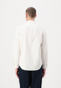 White long-sleeve shirt with a textured fabric, featuring a curved hem and button cuffs. View from the back, showing no visible patterns.