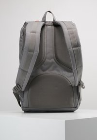 Sac à dos gris avec un panneau arrière en maille rembourrée et texturée et des bretelles ajustables. Présente un design épuré et des boucles latérales doubles.