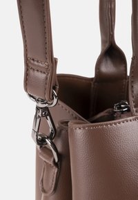 Esquina de bolso de cuero marrón con una correa cosida sujeta con hebillas metálicas plateadas, mostrando superficie texturizada y cremallera dentro del bolso.