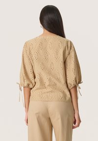 Beige embroidered blouse featuring eyelet patterns, short puffed sleeves with ties, and a relaxed fit. The fabric is textured and lightweight.