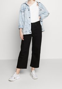 Hellblaue Denimjacke über einem weißen T-Shirt, kombiniert mit schwarzen weit geschnittenen Hosen und weißen Sneakers. Lässiger Streetwear-Look.