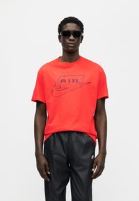 Κόκκινη βαμβακερή μπλούζα με μπλε λογότυπο "AIR" και σήμα Nike, συνδυασμένη με μαύρο αθλητικό παντελόνι. Το μοντέλο φοράει σκούρα γυαλιά ηλίου.