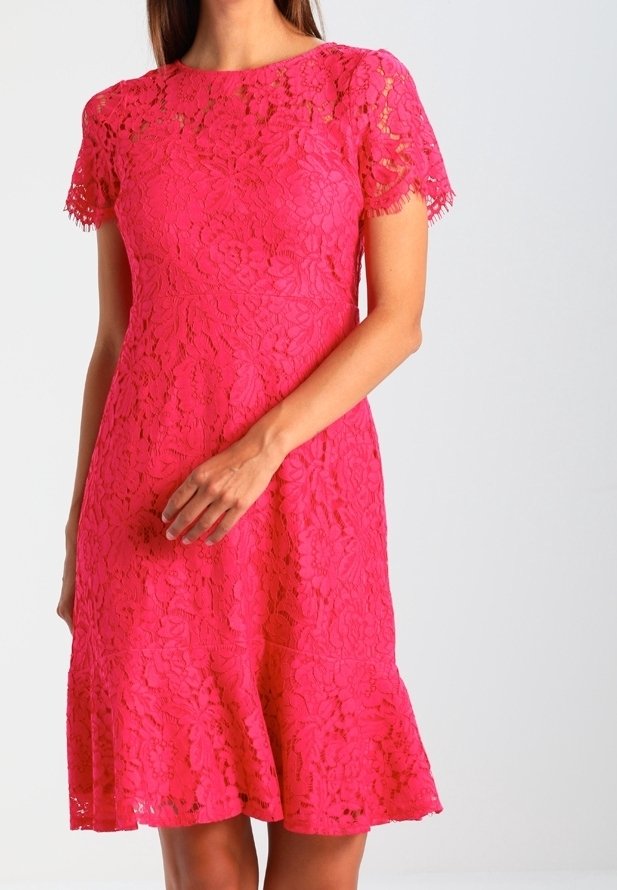 Femme portant une robe en dentelle rose vif avec des manches courtes et un ourlet évasé, debout devant un fond clair uni.