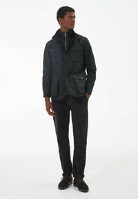 Zwarte lichte jas met vier voorzakken, knoopsluiting en ritskraag. Gecombineerd met donkere jeans en bruine schoenen.