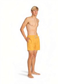 Jaunas nuogas vyras su šviesiais plaukais, dėvintis geltonas maudymosi kelnes, stovi basomis kojomis su rankomis kišenėse prieš baltą foną.