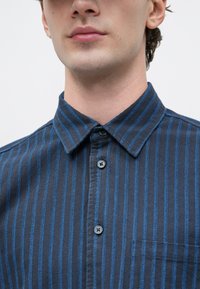 Primer plano de una persona con una camisa azul marino con rayas verticales de color azul claro y botones negros, mostrando el cuello y la parte superior del pecho.