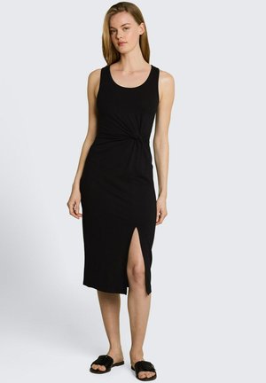 Frau trägt ein ärmelloses schwarzes Midikleid mit seitlichem Schlitz und Knotendetail in der Taille, kombiniert mit schwarzen Slide-Sandalen.