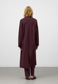 Lange bordeauxrode jas met een rechte snit, verlaagde schouders, zijsplitsen en bijpassende broeken. Glad textuur, minimalistisch design.
