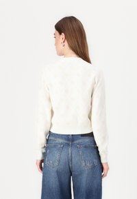 Cream-colored sweater with diamond pattern, ribbed cuffs, and hem. Featuring a round neckline, paired with high-waisted blue jeans.