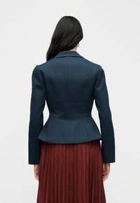Blazer bleu marine sur mesure avec taille peplum, épaules structurées et manches longues, associé à une jupe plissée bordeaux foncé.